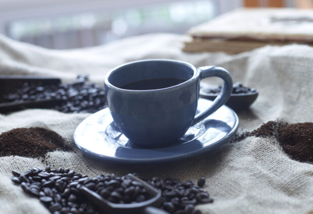 Black Coffee in a Blue Cup with Saucer on a Burlap Material Surface with Coffee Beans and Ground Coffee in an Indoor Setting