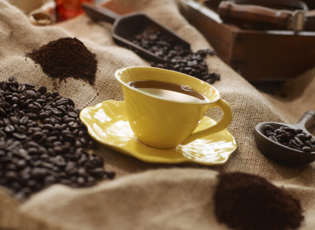 Black Coffee in a Yellow Cup with Saucer on a Burlap Material Surface with Coffee Beans and Ground Coffee in an Indoor Setting