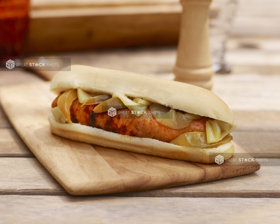 Close Up of a Hot Dog with a Grilled Sausage and Roasted Onions and Peppers on a Wooden Cutting Board on a Wooden Table in an Indoor Setting