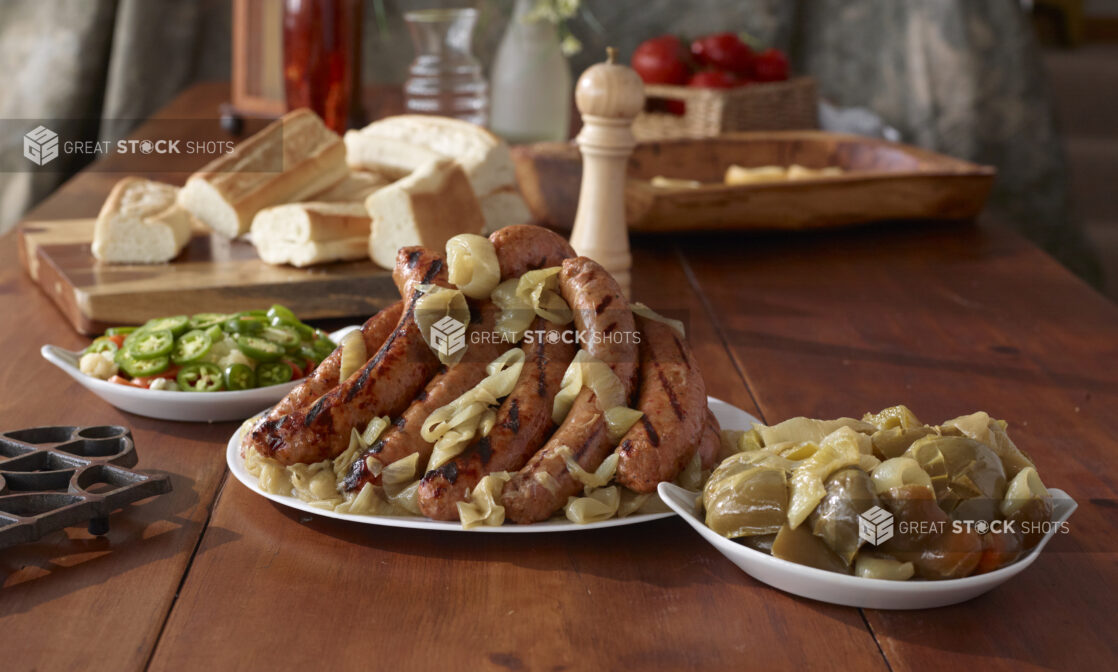 Platter of Grilled Sausage Links and Roasted Onions with Side Dishes of Cooked and Fresh Vegetable Accompaniments and Sliced Baguettes on a Wooden Table Indoors