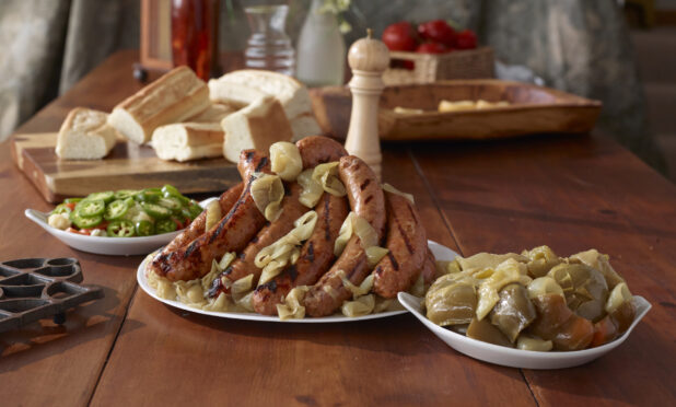 Platter of Grilled Sausage Links and Roasted Onions with Side Dishes of Cooked and Fresh Vegetable Accompaniments and Sliced Baguettes on a Wooden Table Indoors