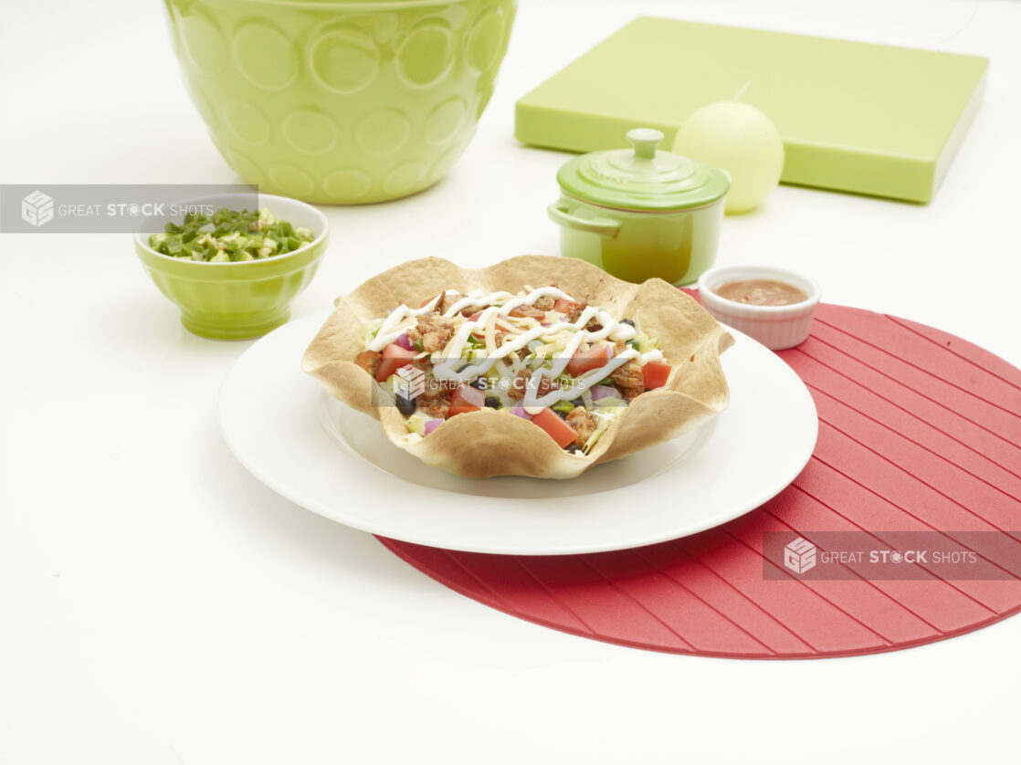 Spicy Grilled Chicken Mexican Taco Salad in a Crispy Tortilla Bowl on a Round White Ceramic Dish on a Red Placemat on a White Table in an Indoor Setting