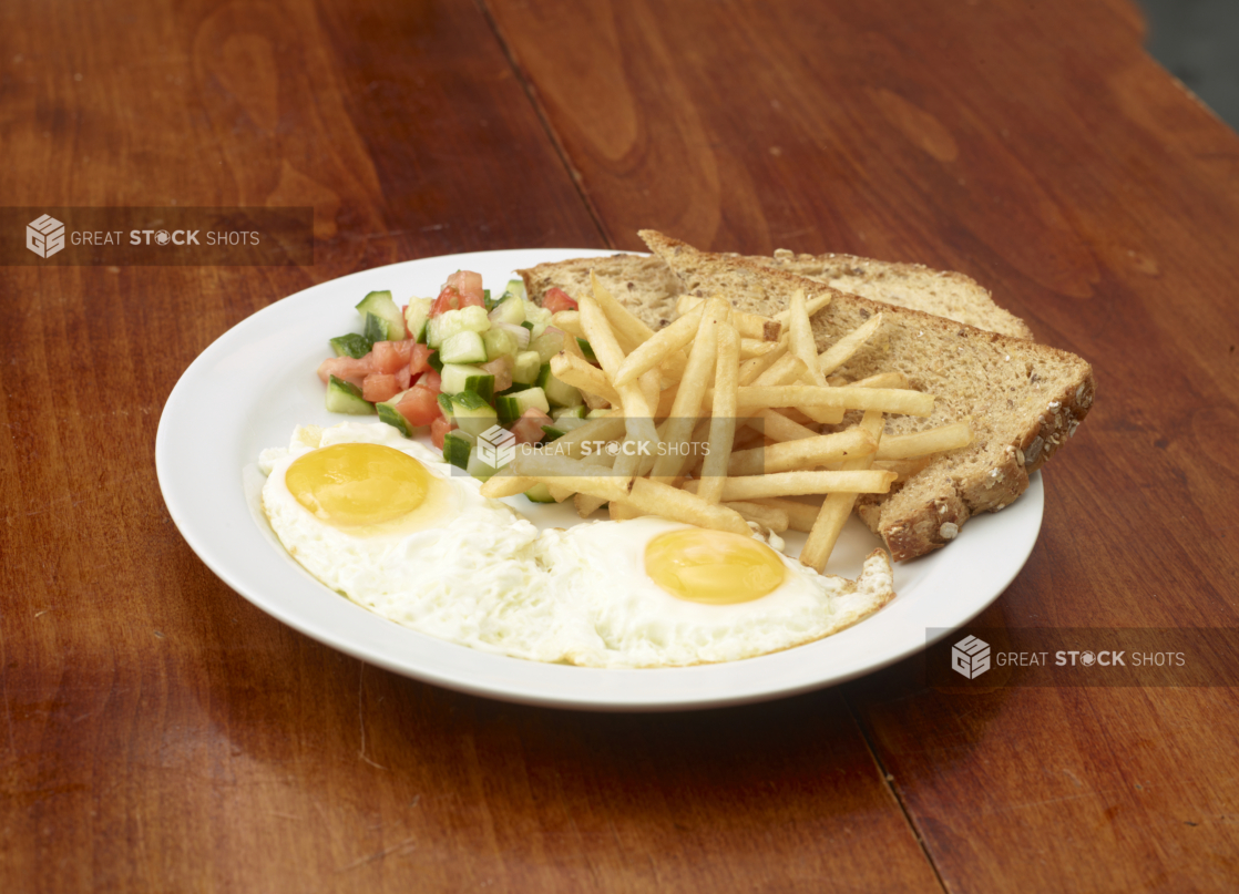 Two eggs sunny side up with french fries, multigrain toast, and chopped tomato and cucumber salad