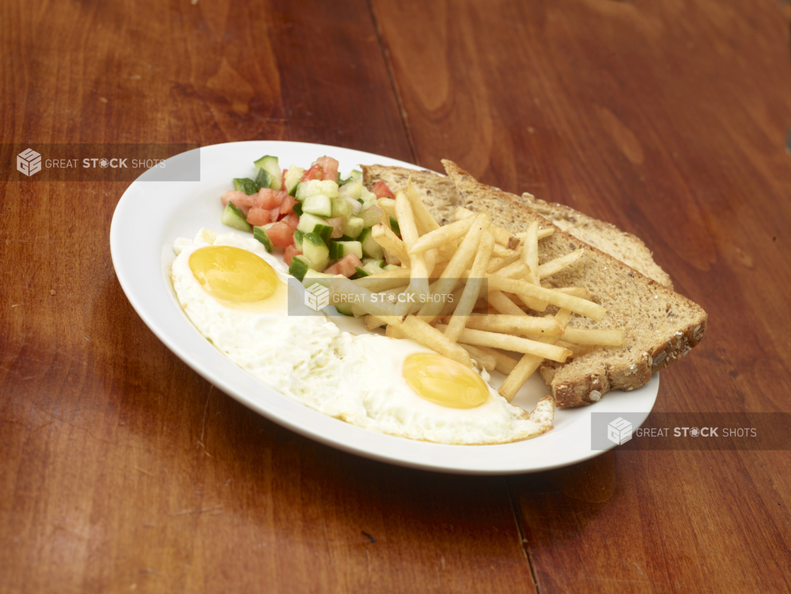 Two eggs sunny side up with french fries, multigrain toast, and chopped tomato and cucumber salad