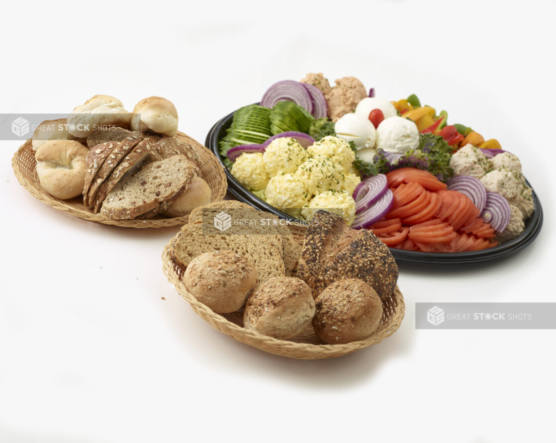 Dairy Platter with tuna salad, egg salad, salmon salad and cream cheese in mounds with sliced tomato, sliced cucumber, sliced red onions and slices of bell pepper all on a black catering tray with 2 small baskets of bread and buns