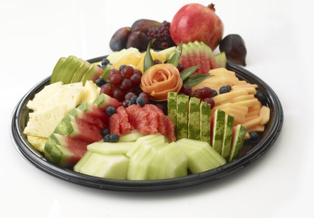 Fruit tray of sliced honey dew melon, watermelon, cantaloupe, pineapple, oranges, red grapes and blueberries all on a round black catering tray with whole pomegranate, figs and grapes in the background