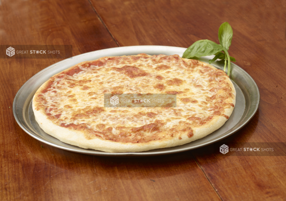 A Whole Cheese Pizza with Fresh Basil Garnish on a Silver Platter on a Wooden Table