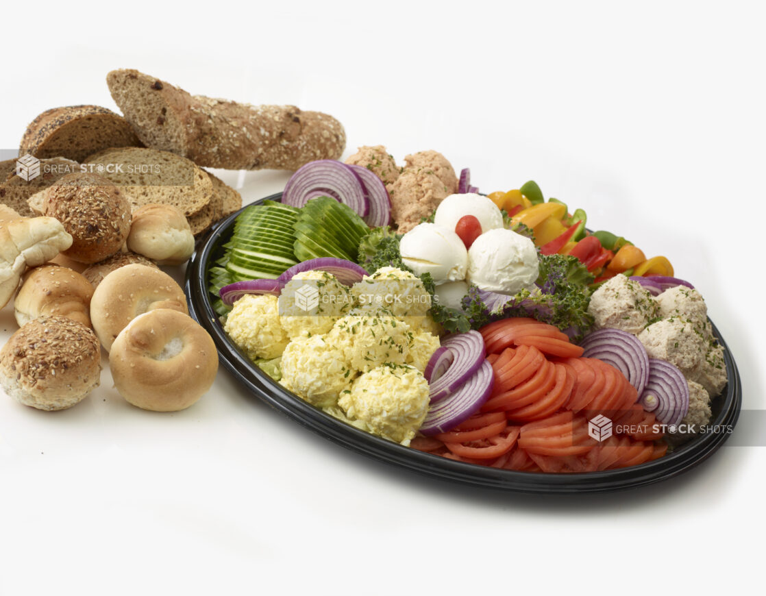 Dairy Platter with tuna salad, egg salad, salmon salad and cream cheese in mounds with sliced tomato, sliced cucumber, sliced red onions and slices of bell pepper all on a black catering tray with breads, rolls and bagels in the background