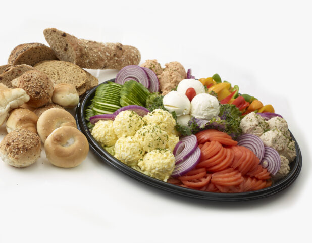 Dairy Platter with tuna salad, egg salad, salmon salad and cream cheese in mounds with sliced tomato, sliced cucumber, sliced red onions and slices of bell pepper all on a black catering tray with breads, rolls and bagels in the background