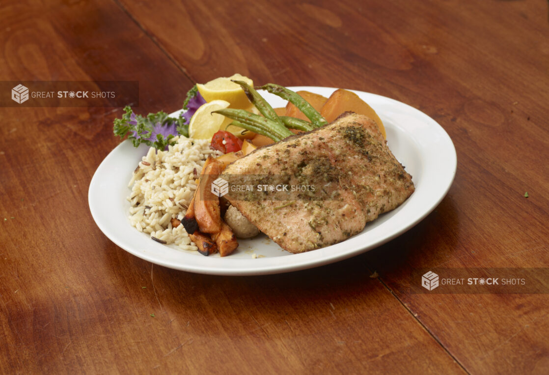 Grilled salmon with rice pilaf, roasted potatoes, carrots, green beans and slices of lemons on a round white plate on a wooden background