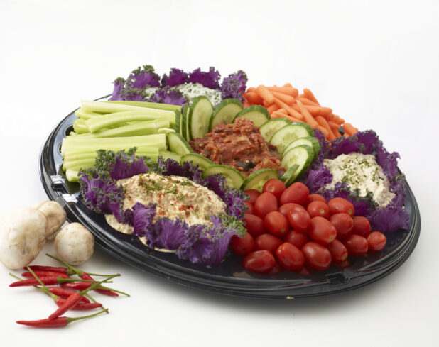 Vegetable and dip platter with 4 dips and fresh grape tomatoes, baby carrots, celery and cucumber on a round black catering tray with fresh mushrooms and red chilis beside the platter