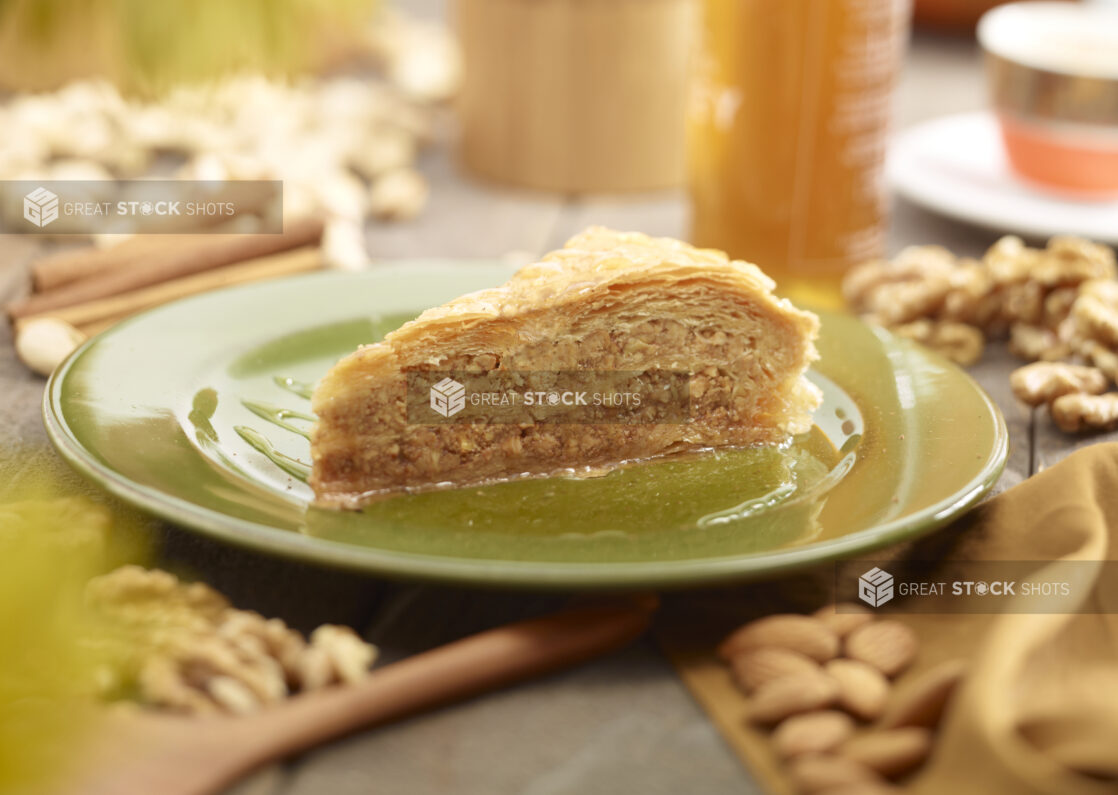 A slice of baklava on a green side plate surrounded by nuts and honey, close up view