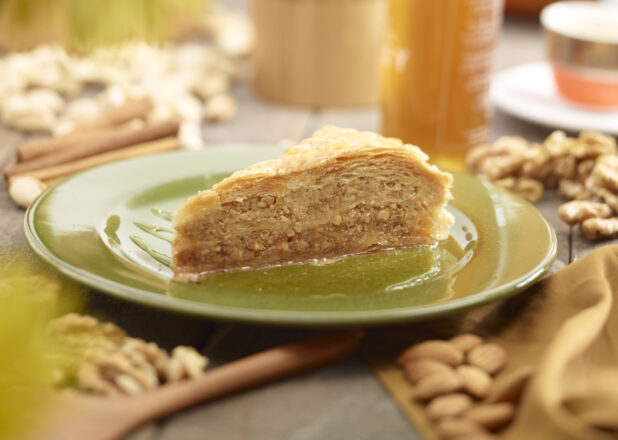 A slice of baklava on a green side plate surrounded by nuts and honey, close up view