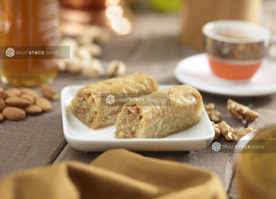 2 pieces of baklava on a square white side plate with nuts, honey, napkin and middle eastern coffee cup in the surrounding background