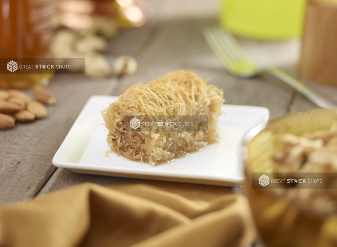 Baklava on a square white side plate with nuts, honey, napkin in the surrounding background