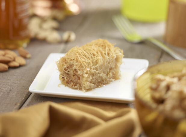 Baklava on a square white side plate with nuts, honey, napkin in the surrounding background