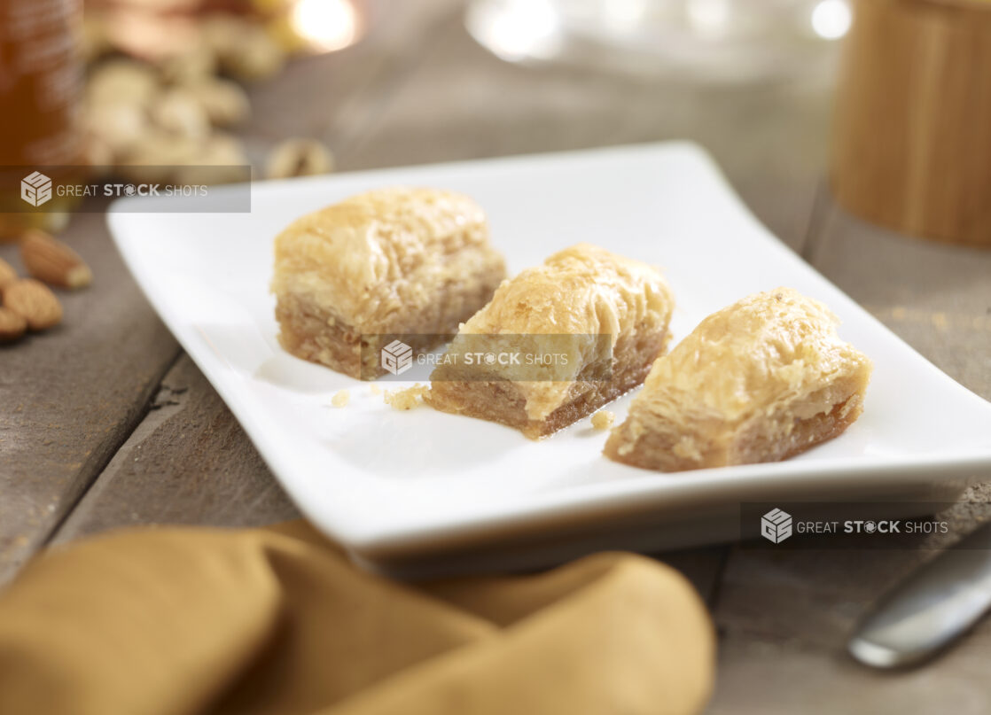 3 pieces of baklava on a square white side plate with nuts, honey, napkin in the surrounding background