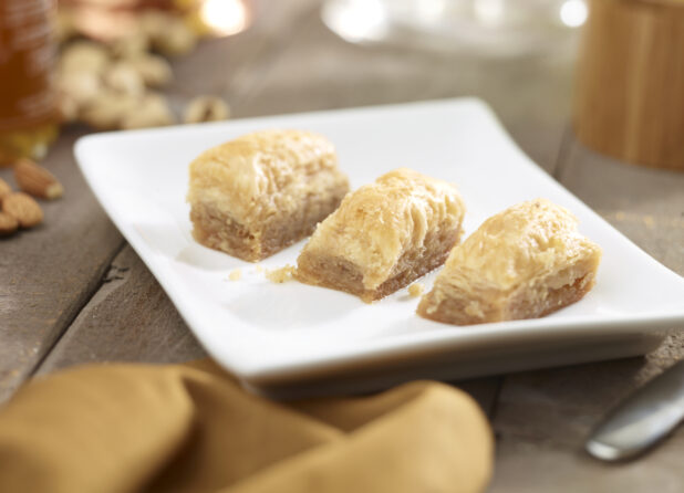 3 pieces of baklava on a square white side plate with nuts, honey, napkin in the surrounding background