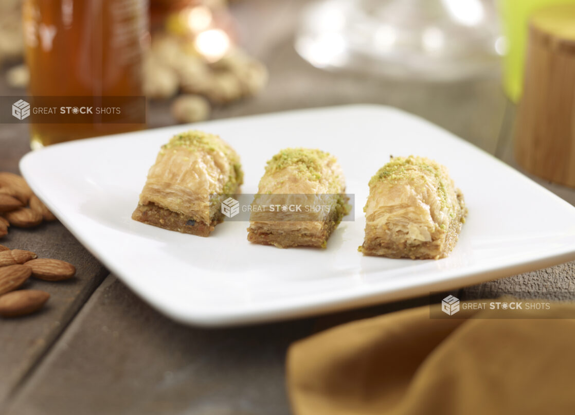 3 pieces of baklava on a square white side plate with nuts, honey, napkin in the surrounding background