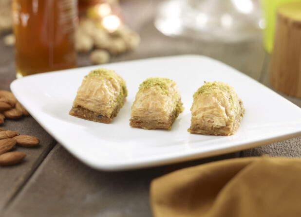 3 pieces of baklava on a square white side plate with nuts, honey, napkin in the surrounding background