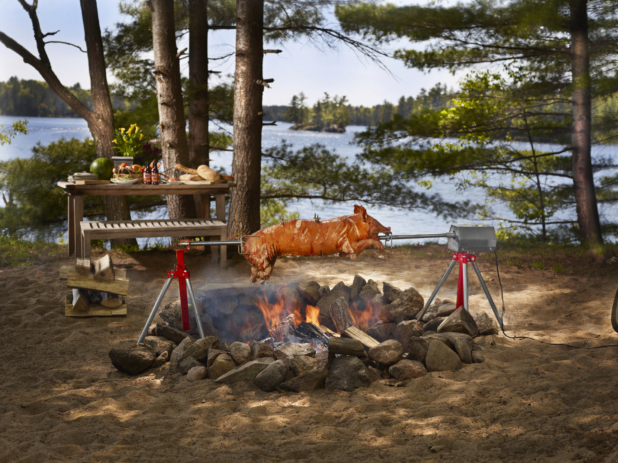 Pig roasting on a spit over a campfire in a wooded area with lake in background