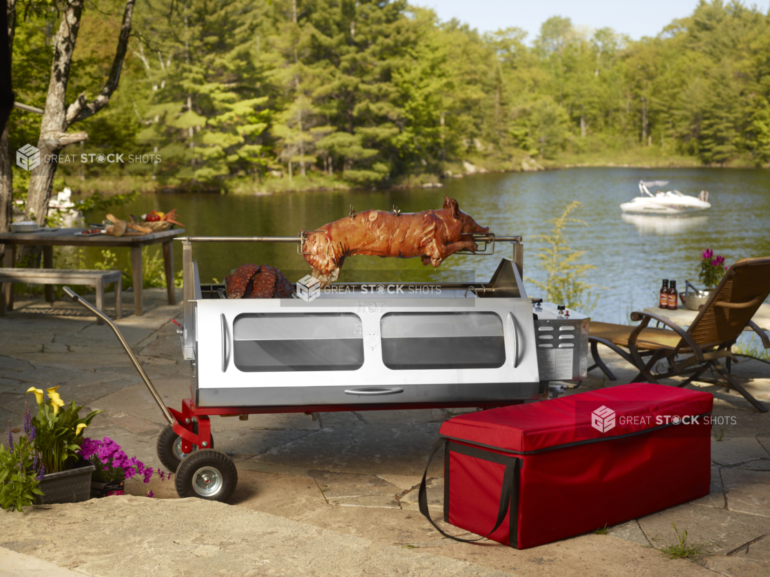 Whole pig roasting on a spit over a propane roaster on a stone terrace with pond and woods in background