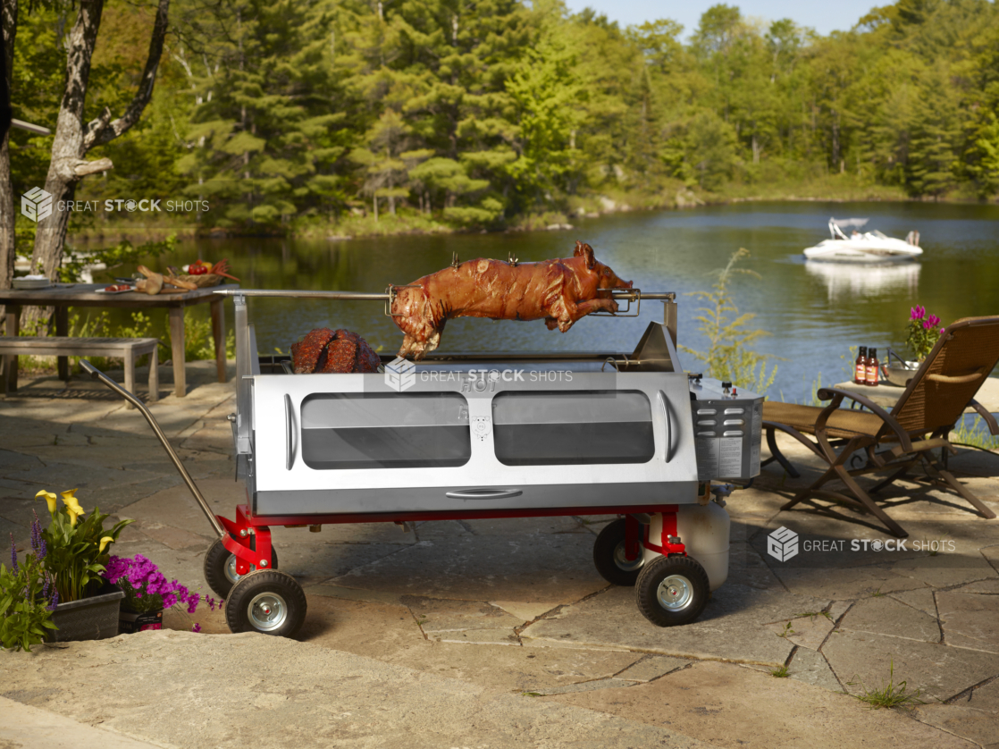 Whole pig roasting on a spit over a propane roaster on a stone terrace with pond and woods in background