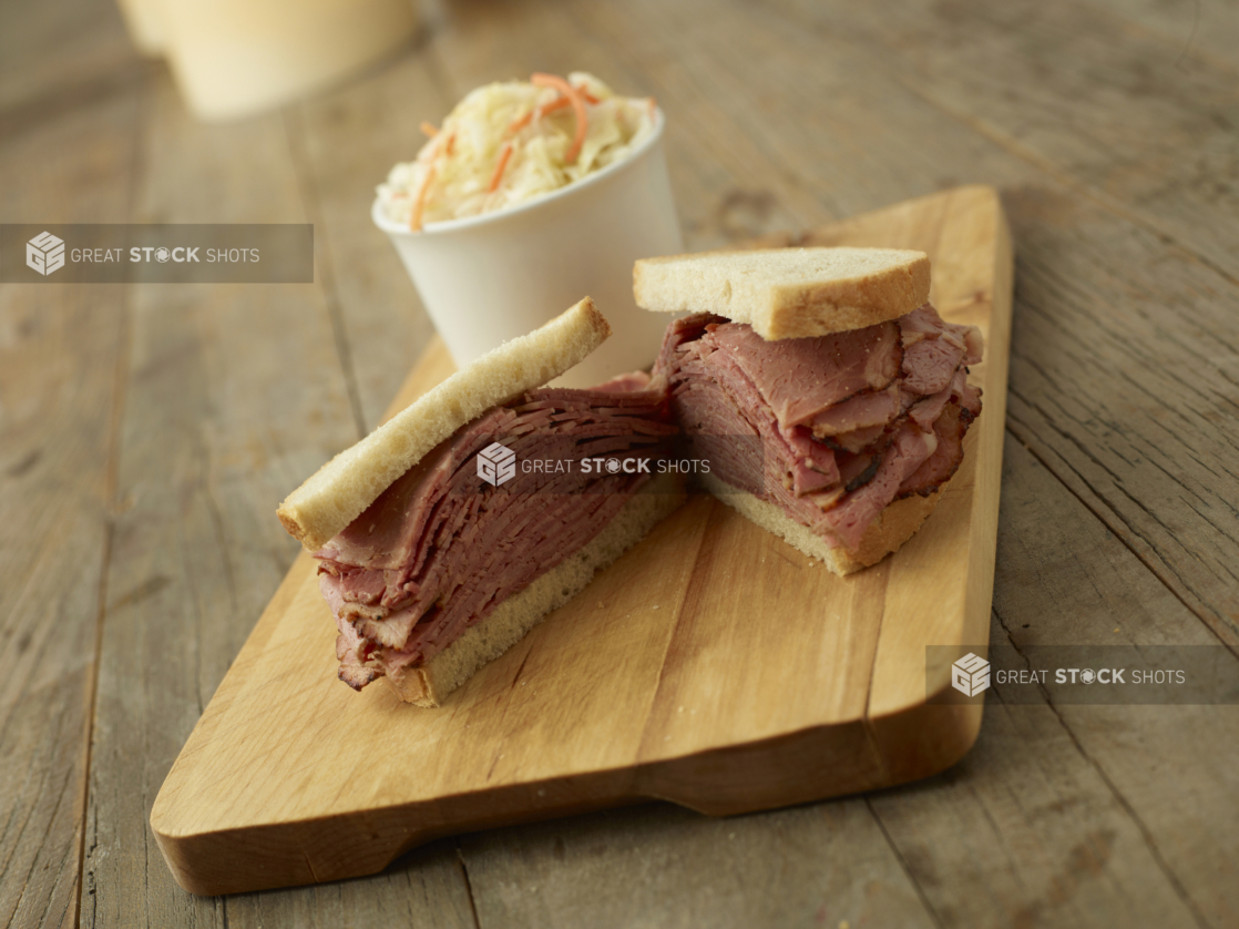 A Thick Deli-Style Pastrami Sandwich on White Rye Bread Cut Open on a Wooden Cutting Board with a Cup of Coleslaw in the Background