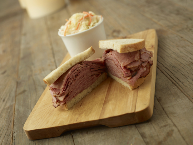 A Thick Deli-Style Pastrami Sandwich on White Rye Bread Cut Open on a Wooden Cutting Board with a Cup of Coleslaw in the Background