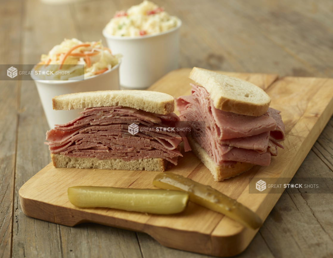 A Thick Deli-Style Pastrami Sandwich on White Rye Bread Cut Open on a Wooden Cutting Board with Pickle Spears and Cups of Coleslaw in the Background