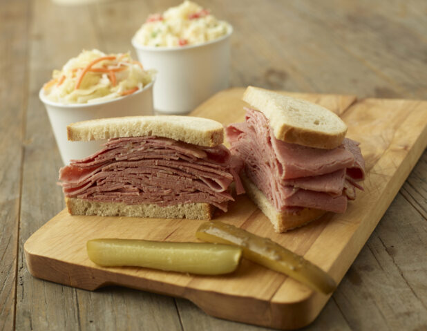 A Thick Deli-Style Pastrami Sandwich on White Rye Bread Cut Open on a Wooden Cutting Board with Pickle Spears and Cups of Coleslaw in the Background