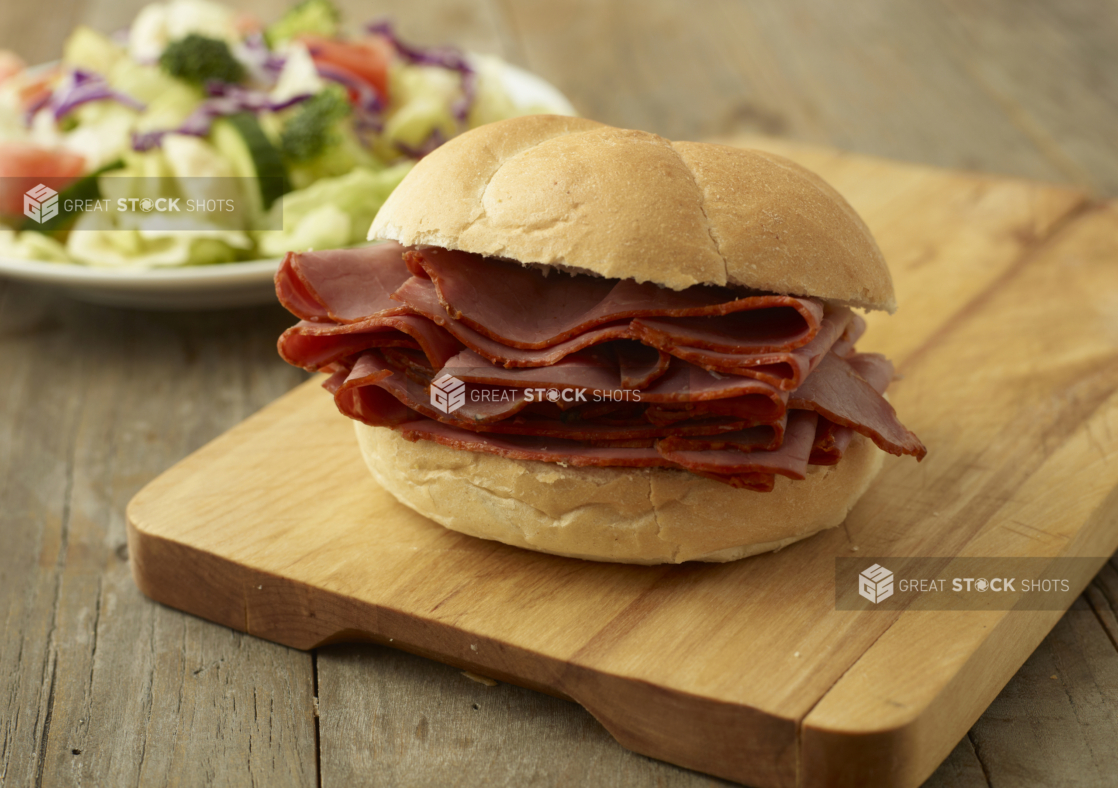 Thick-Cut Pastrami Deli Meat Piled High in a Kaiser Bun on a Wooden Cutting Board with a Garden Salad in the Background