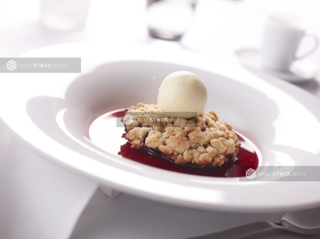 Cherry Crumble with a Scoop of French Vanilla Bean Ice Cream and Berry Coulis in a White Ceramic Bowl on a White Table Cloth Surface