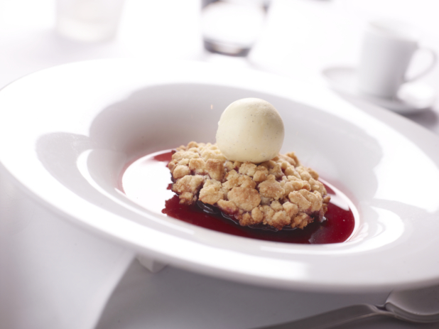 Cherry Crumble with a Scoop of French Vanilla Bean Ice Cream and Berry Coulis in a White Ceramic Bowl on a White Table Cloth Surface