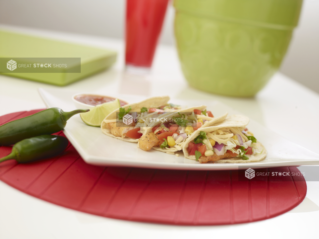 3 fish tacos and a slice of lime on a square white plate, jalapenos beside