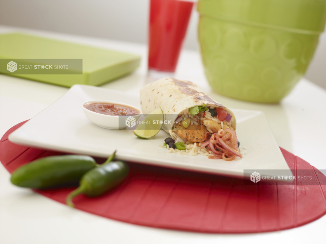 Shrimp burrito with a slice of lime and a ramekin of red salsa on a square white plate, whole jalapenos on the side