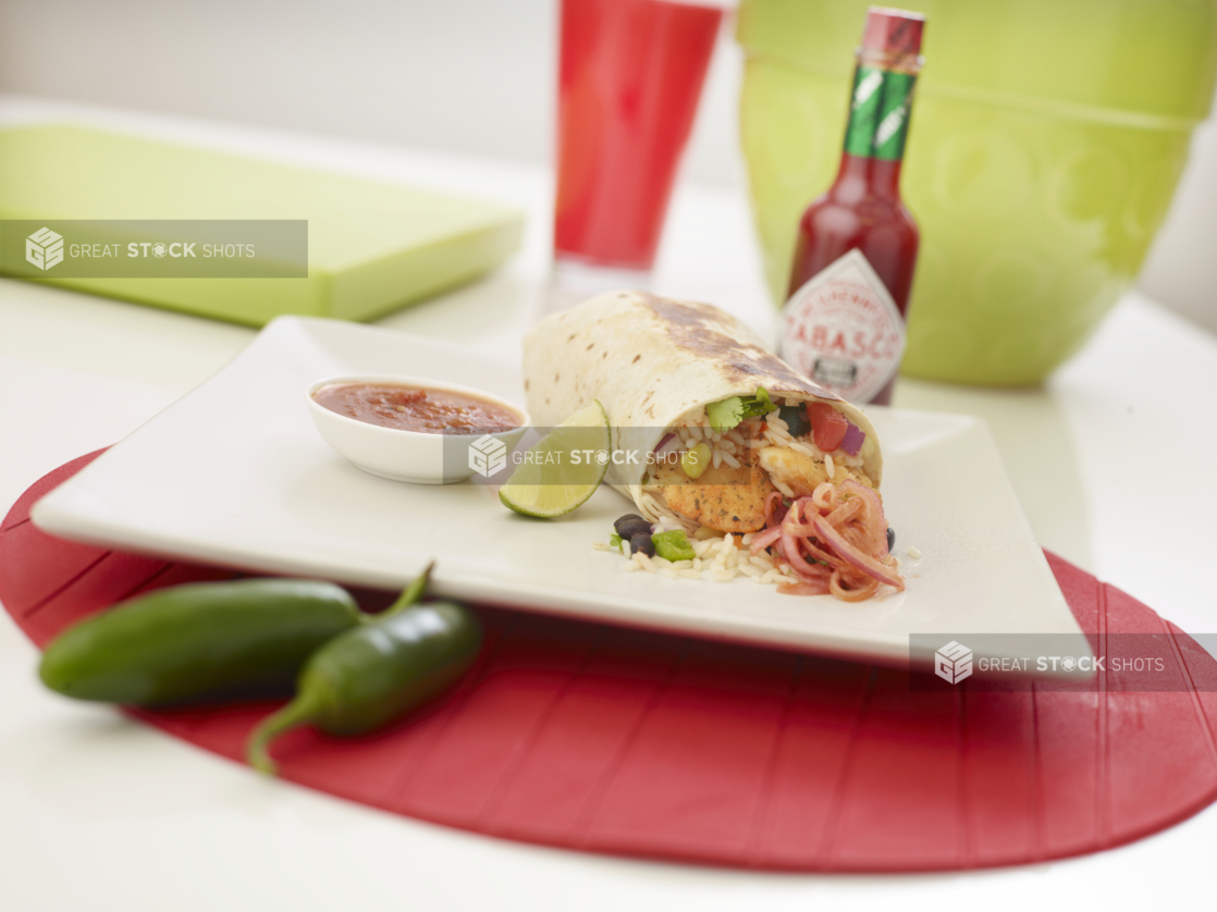 Shrimp burrito with a slice of lime and a ramekin of red salsa on a square white plate