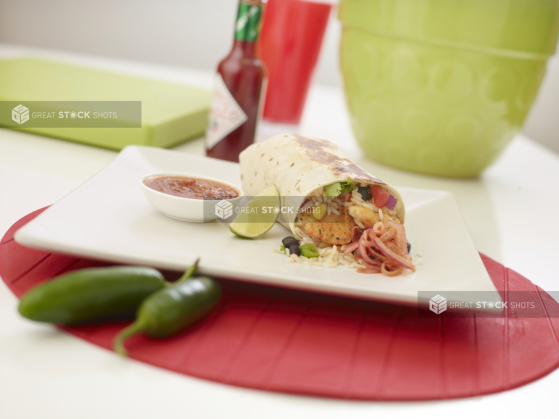 Shrimp burrito with a slice of lime and a ramekin of red salsa on a square white plate