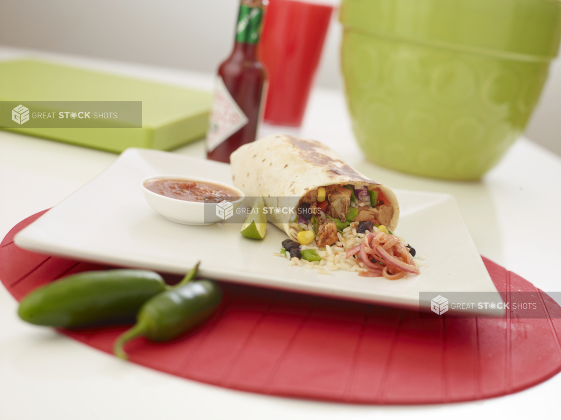 Shrimp burrito with a slice of lime and a ramekin of red salsa on a square white plate, Tabasco sauce in background