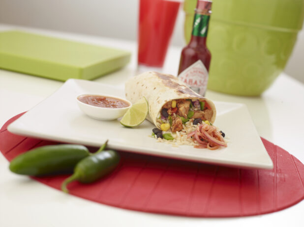 Shrimp burrito with a slice of lime and a ramekin of red salsa on a square white plate, Tabasco sauce in background