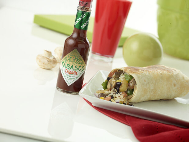 Steak and mushroom burrito with a slice of lime and a ramekin of red salsa on a square white plate beside a bottle of Tabasco sauce