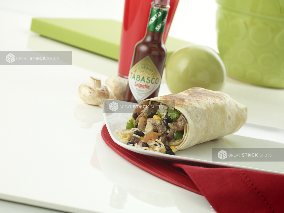 Steak burrito with a slice of lime and a ramekin of red salsa on a square white plate beside a bottle of Tabasco sauce