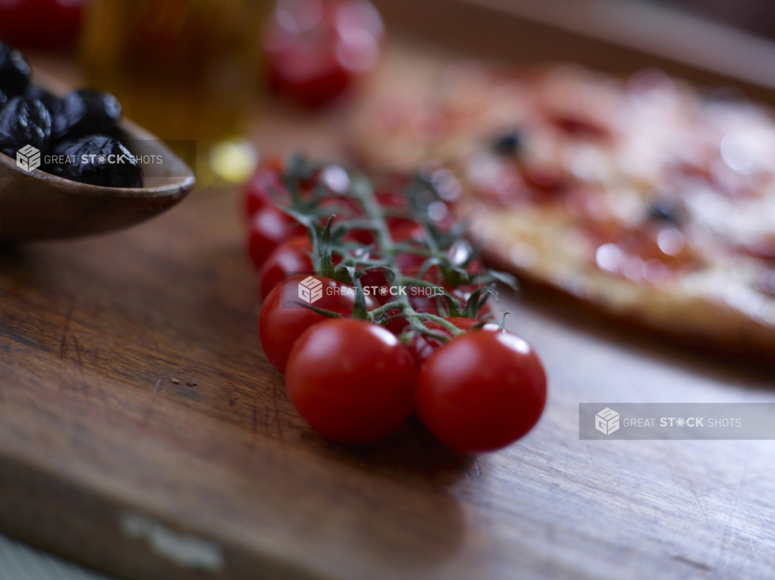 Cherry tomatoes on the vine on a wood board with black olives to the side and a pizza in the background