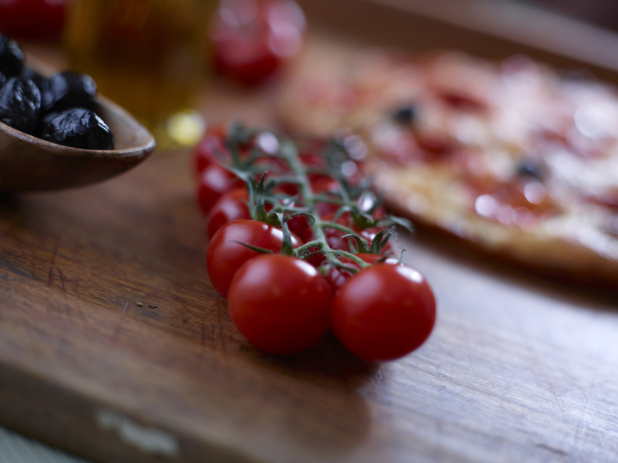 Cherry tomatoes on the vine on a wood board with black olives to the side and a pizza in the background