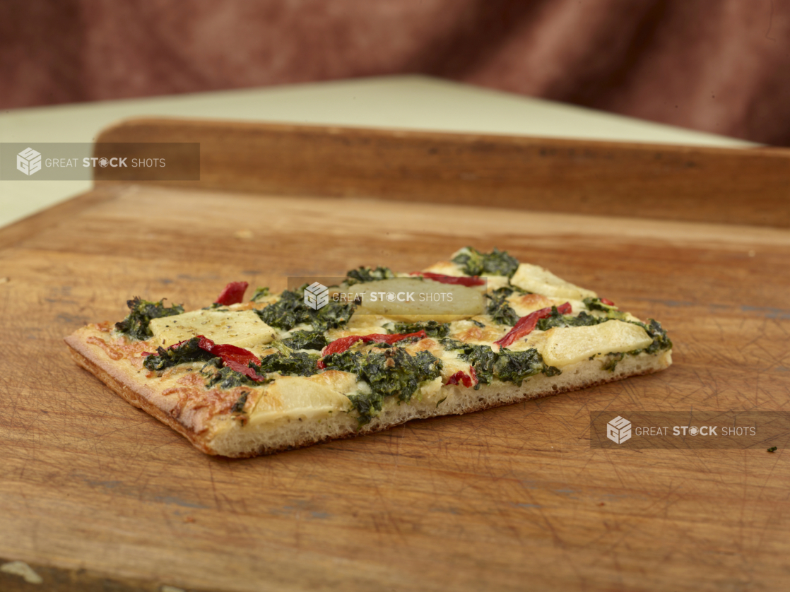 Close Up of a Slice of Sicilian-Style Rectangular Pizza with Potato, Spinach and Roasted Red Pepper Toppings on a Wooden Board