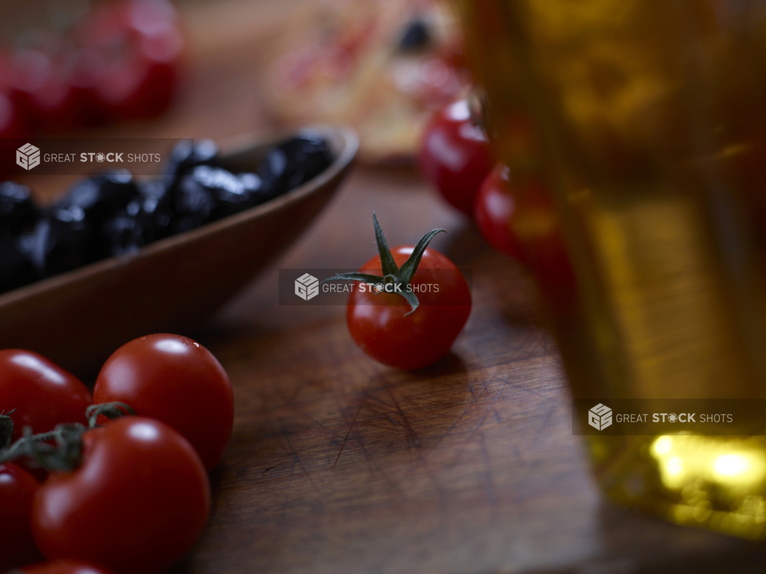 Loose cherry tomatoes on a wood cutting board with a bottle of olive oil in the foreground and a dish of black olives in the background, close-up, low angle