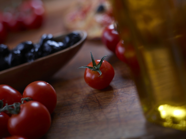 Loose cherry tomatoes on a wood cutting board with a bottle of olive oil in the foreground and a dish of black olives in the background, close-up, low angle