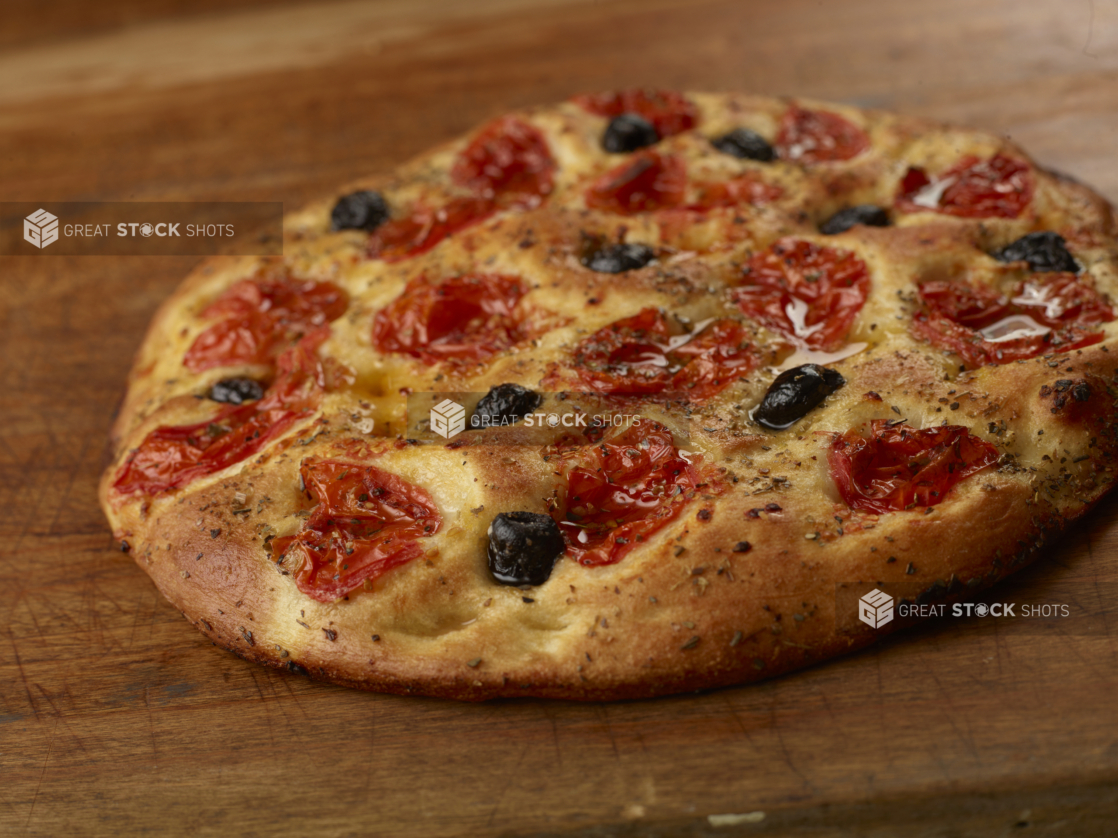 Round herbed cherry tomato and black olive flatbread on a wooden cutting board, close-up