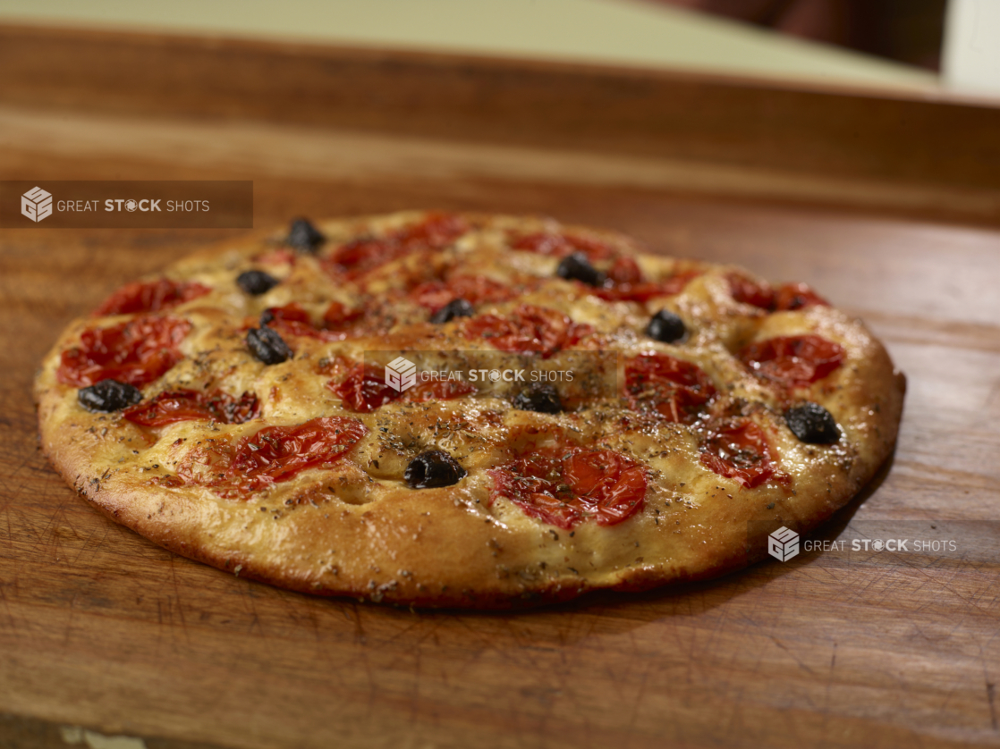 Round herbed cherry tomato and black olive flatbread on a wooden cutting board, close-up