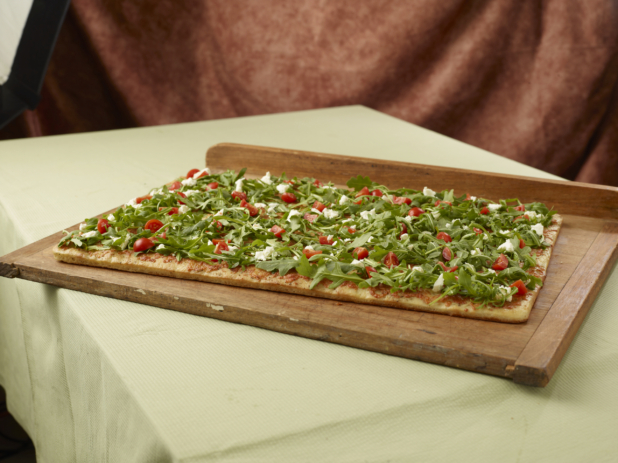 Large rectangular focaccia with arugula, sundried tomato and crumbled white cheese on a wood board, tablecloth